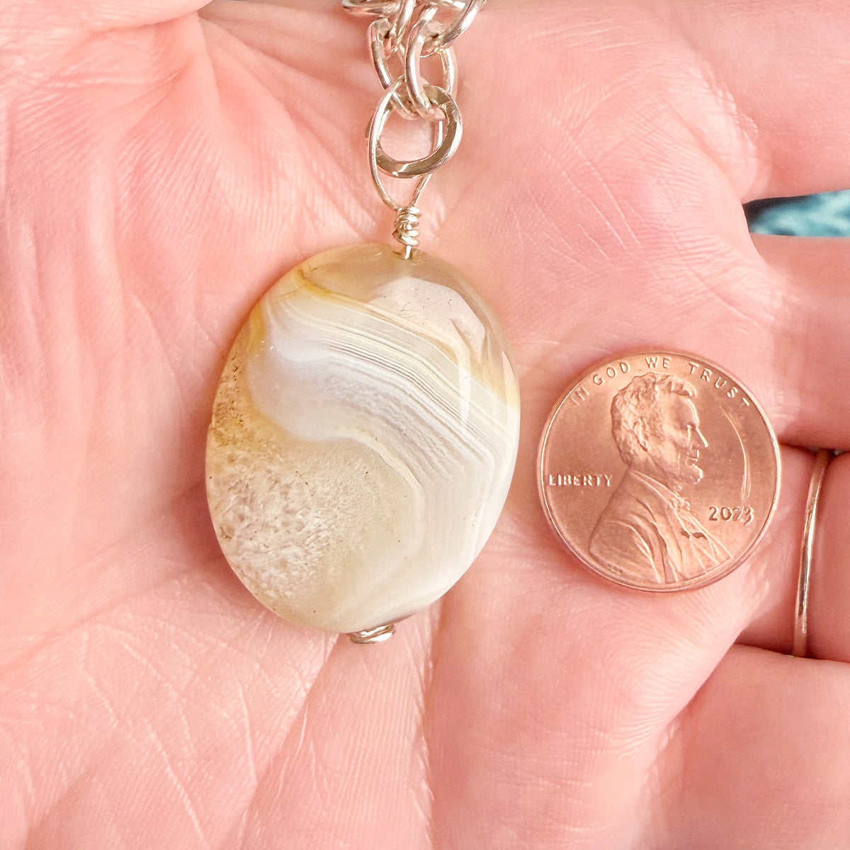 An oval pendant with a white, cream, and light brown agate-like band pattern, hanging from a silver chain and held in a person's palm next to a US penny for scale.