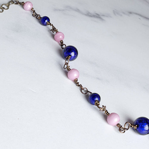 A beaded chain necklace featuring alternating glossy dark blue and matte light pink glass beads, connected by antique bronze-toned metal links, displayed diagonally on a white marble surface.