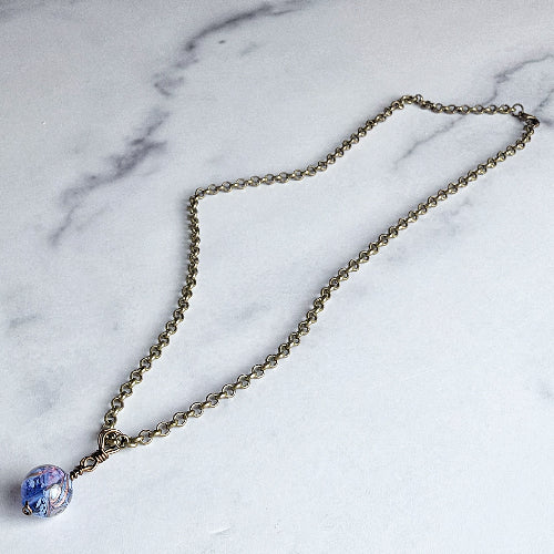 An antique bronze chain necklace with a round glass pendant featuring blue and purple swirls, displayed on a white marble background.