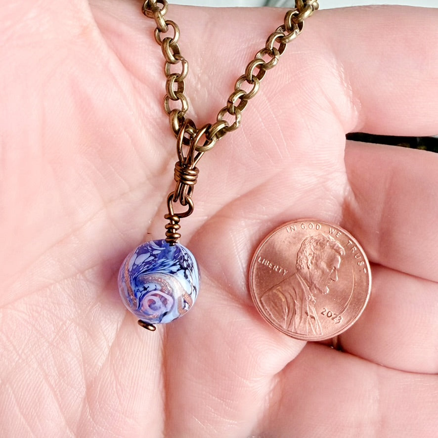 A close-up of a hand holding a bronze chain necklace with a blue and white marbled glass bead pendant, shown next to a U.S.