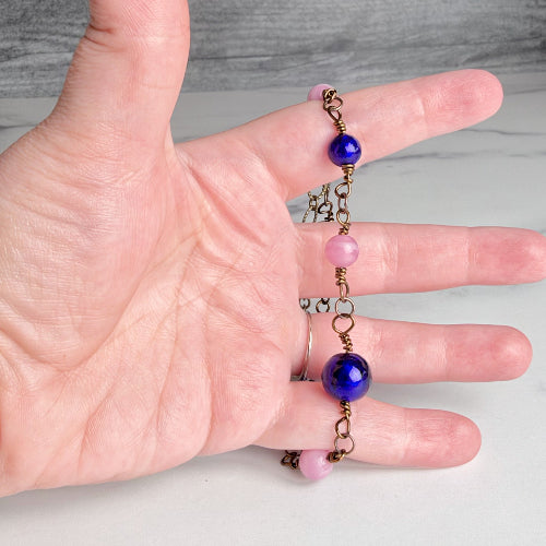 A close-up of a hand holding a delicate bracelet with bronze-toned chain links, pink round beads, and two glossy cobalt blue round beads.
