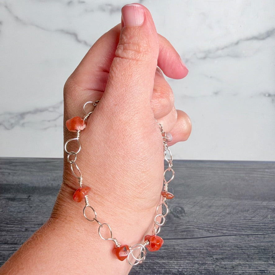 A hand wearing a delicate silver chain bracelet with small orange stone beads, set against a gray wood-grain surface and a white marble background.