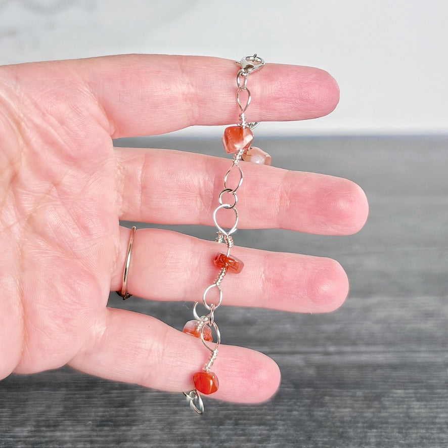 A silver chain bracelet with small orange-red translucent beads, held in a person's hand to show scale against a gray and white background.