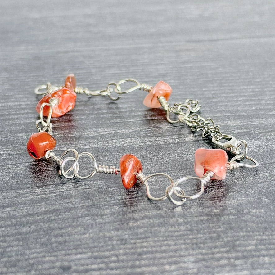 A handmade wire-wrapped bracelet featuring polished orange-red stone beads and silver-toned metal links, displayed on a dark gray wood-textured surface.