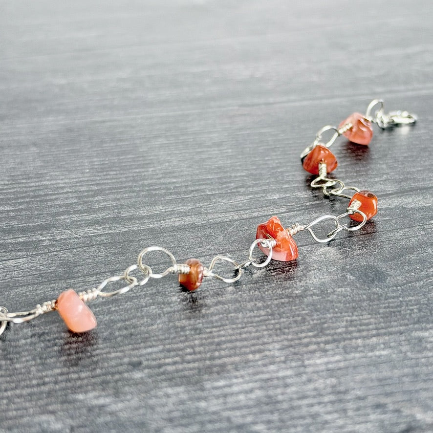A delicate silver wire chain bracelet featuring small, irregular orange-red gemstone beads and a larger pale pink stone bead, displayed on a dark gray wood-textured surface.