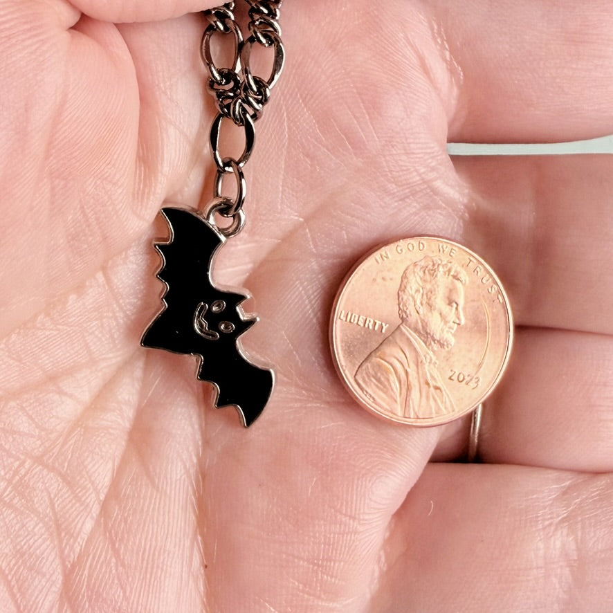 A close-up of a hand holding a black bat-shaped pendant necklace next to a U.S. penny for scale.
