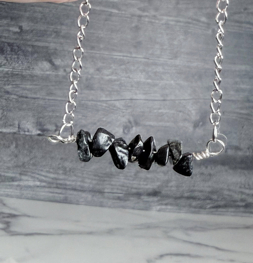 A silver chain necklace featuring a horizontal bar of polished black stone chips, displayed against a gray wood-grain background.