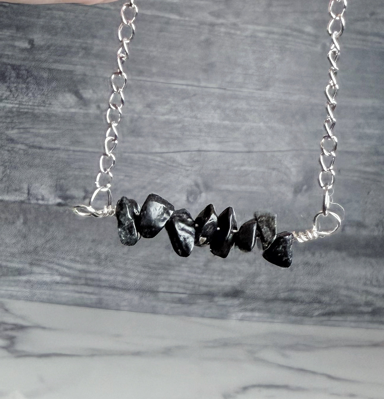 A silver chain necklace featuring a horizontal bar of polished black stone chips, displayed against a gray wood-grain background.