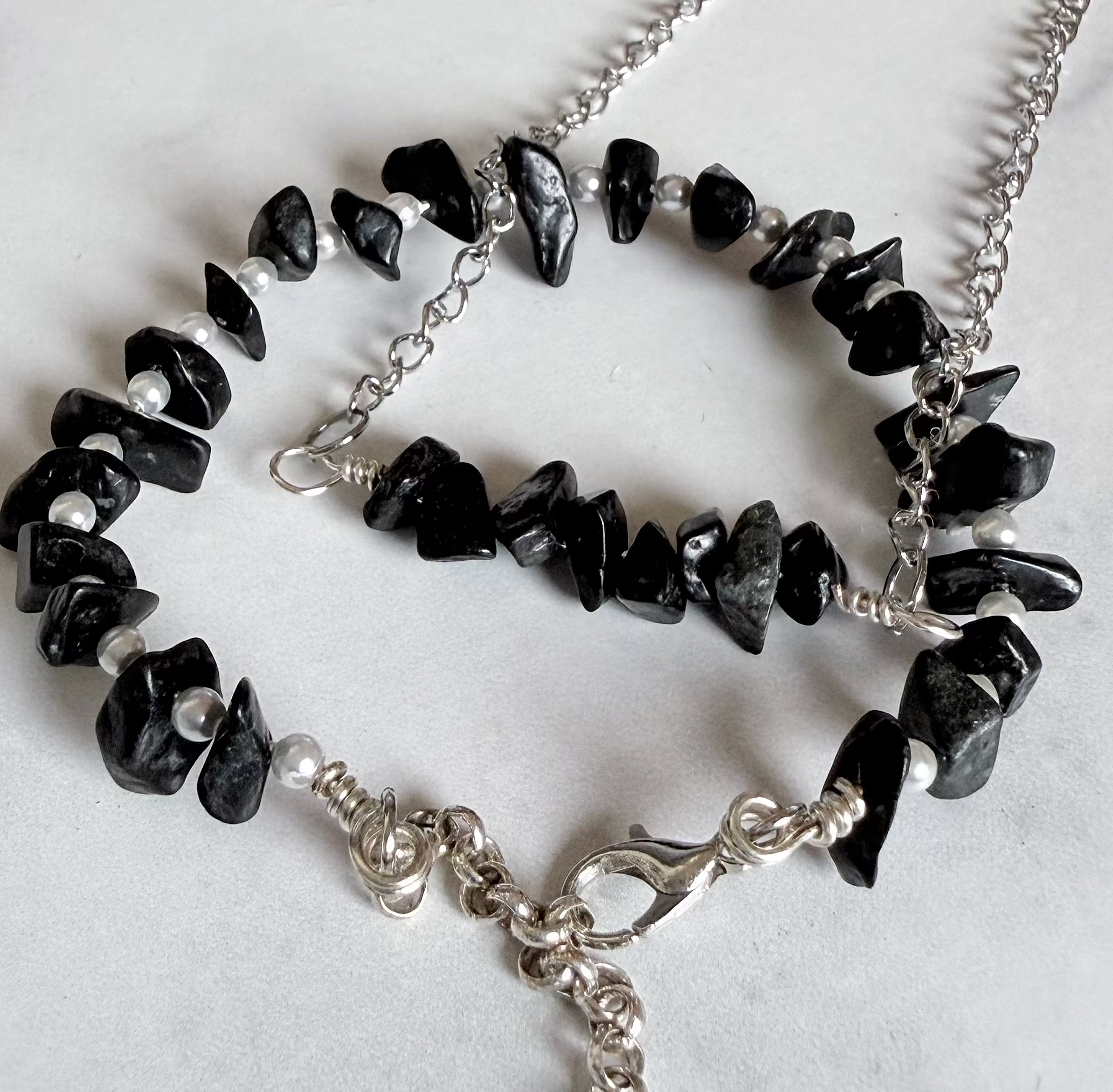 A silver chain necklace and bracelet set featuring polished black chip stones, displayed on a light gray marble surface.