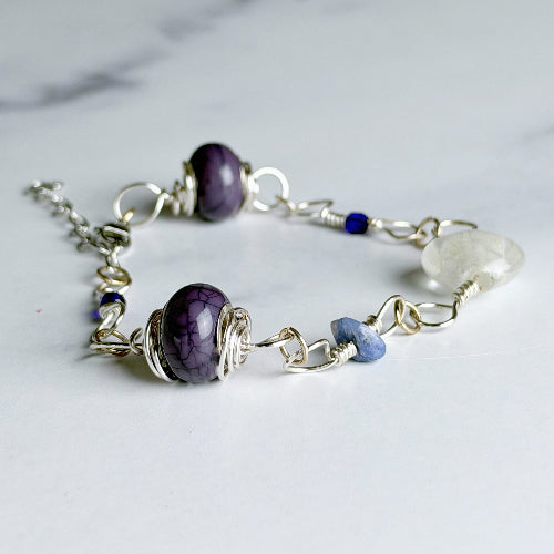 A handmade wire-wrapped bracelet featuring a large purple marbled bead, a smaller blue bead, a clear quartz crystal, and silver chain links, displayed on a light marble surface.