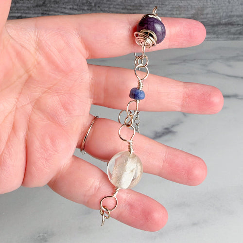 A handmade beaded keychain or charm held in a person's hand. The keychain features a large purple amethyst bead at the top, a small blue bead, several silver wire-wrapped loops, a clear quartz crystal bead, and a small silver ring.