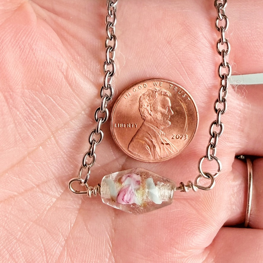 A close-up of a silver-toned chain necklace draped across a person's palm, with a penny placed below the chain for scale and a small clear bead containing pink and white inclusions at the bottom.