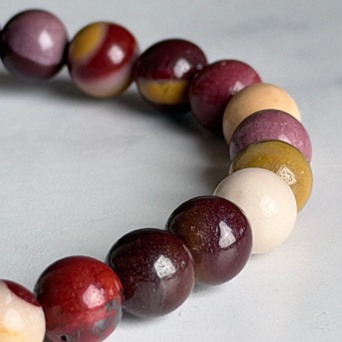 A close-up of a beaded bracelet made of polished round gemstone beads in maroon, cream, yellow, and purple tones, arranged in a curve against a light gray background.