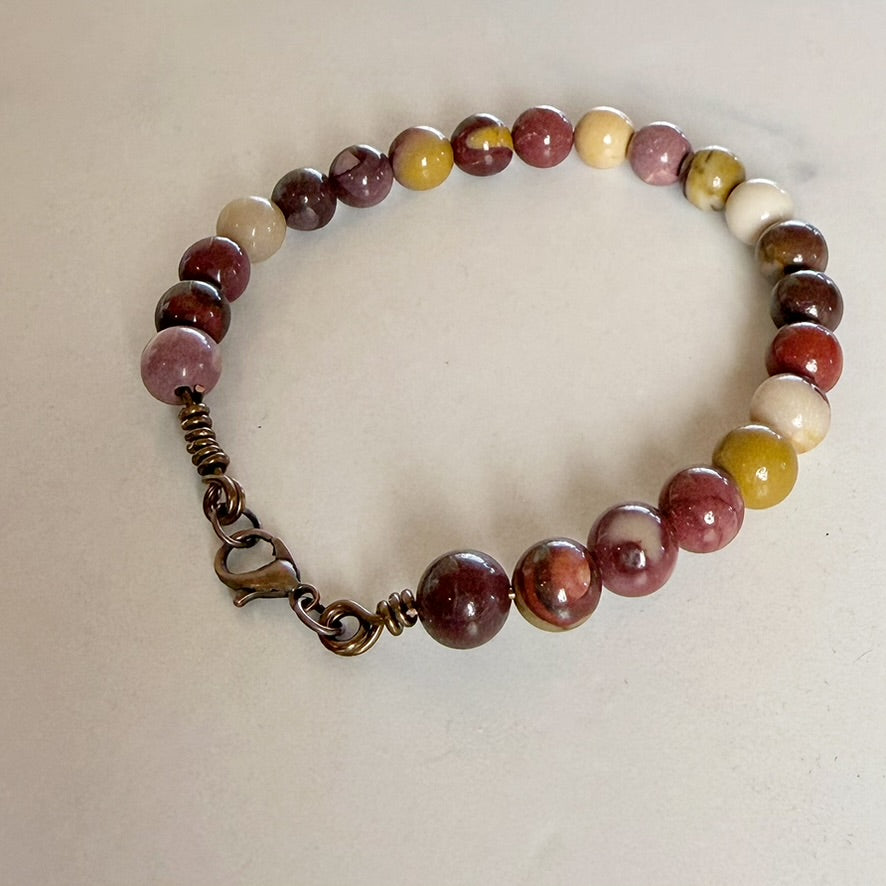 A beaded bracelet made of polished round gemstone beads in shades of burgundy, pink, cream, and olive green, featuring an antique bronze wire-wrapped clasp and lobster clasp, displayed on a light beige surface.