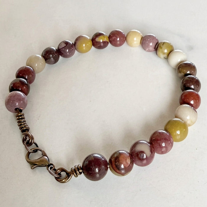 A beaded bracelet made of polished round gemstone beads in shades of burgundy, pink, cream, and olive green, featuring an antique bronze wire-wrapped clasp and lobster clasp, displayed on a light beige surface.