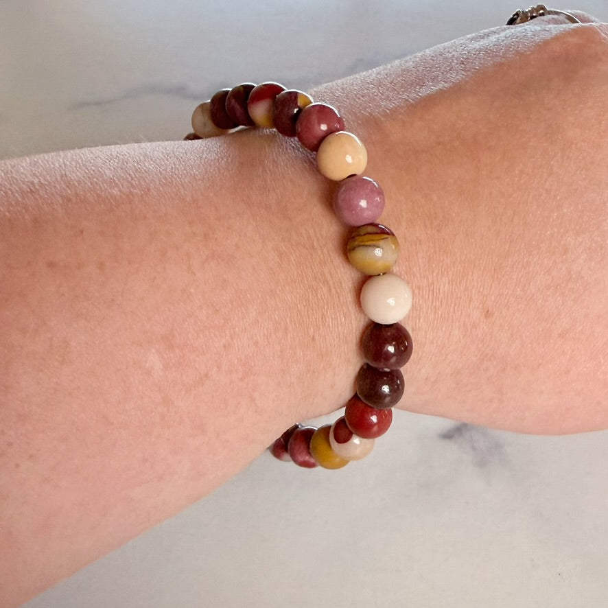 A beaded stretch bracelet worn on a person's wrist, featuring polished round beads in shades of burgundy, cream, pink, and brown.