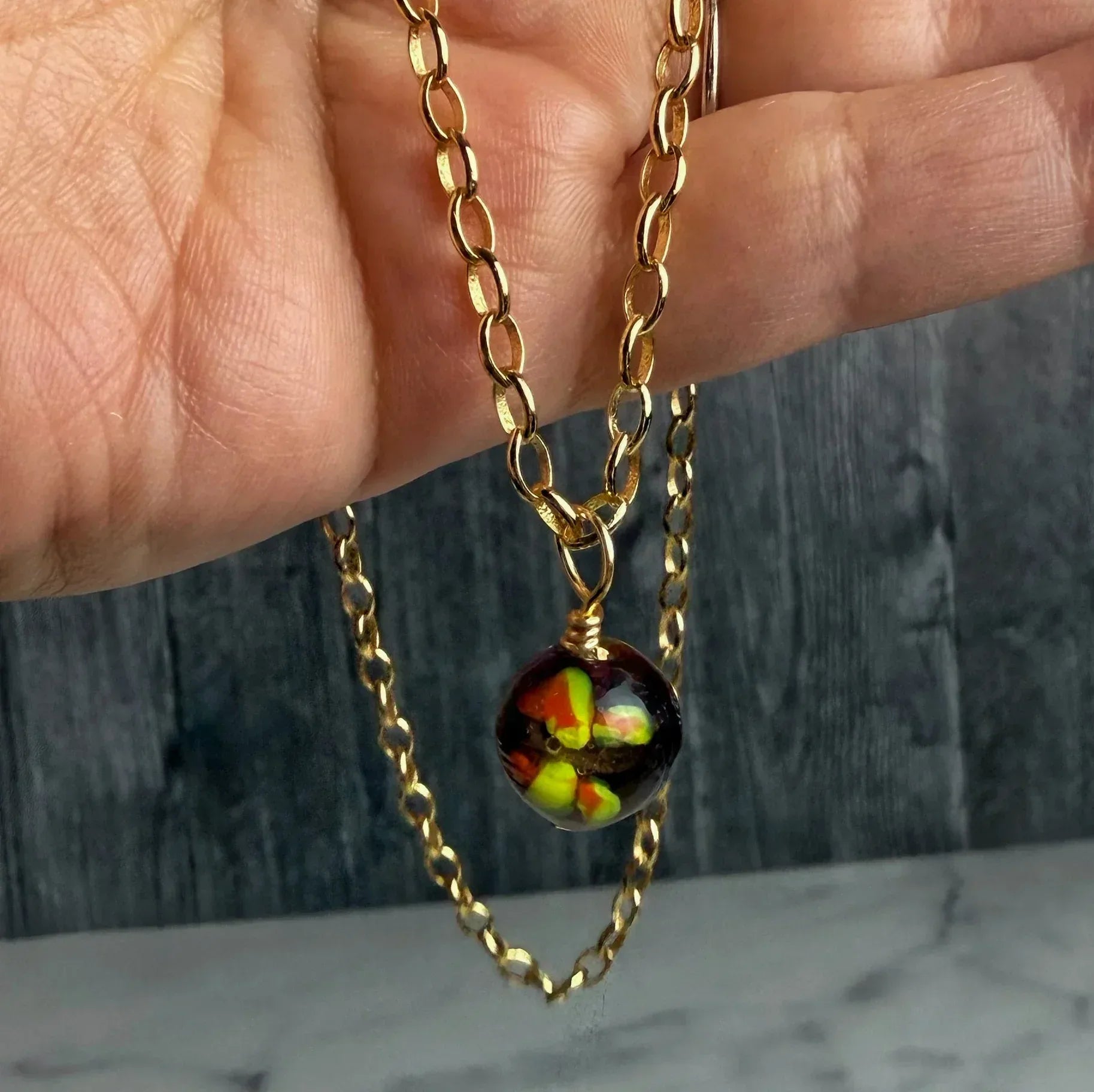 Gold chain necklace with a colorful pendant held by a hand against a wooden background