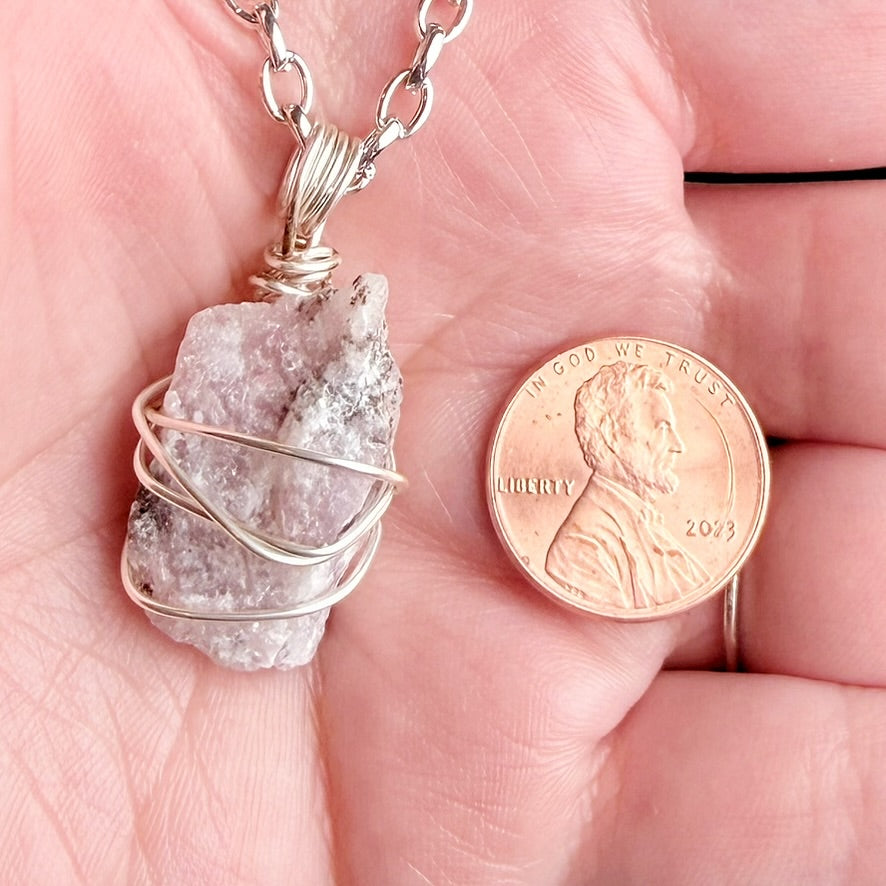 A raw, rough lavender quartz crystal pendant wrapped in silver wire, held in a person's hand next to a US penny for scale.