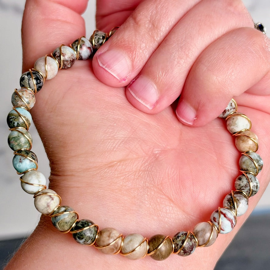 A close-up of a hand holding a beaded stretch bracelet made of polished round stones in earthy beige, brown, white, and green marbled patterns, set in gold-toned metal.