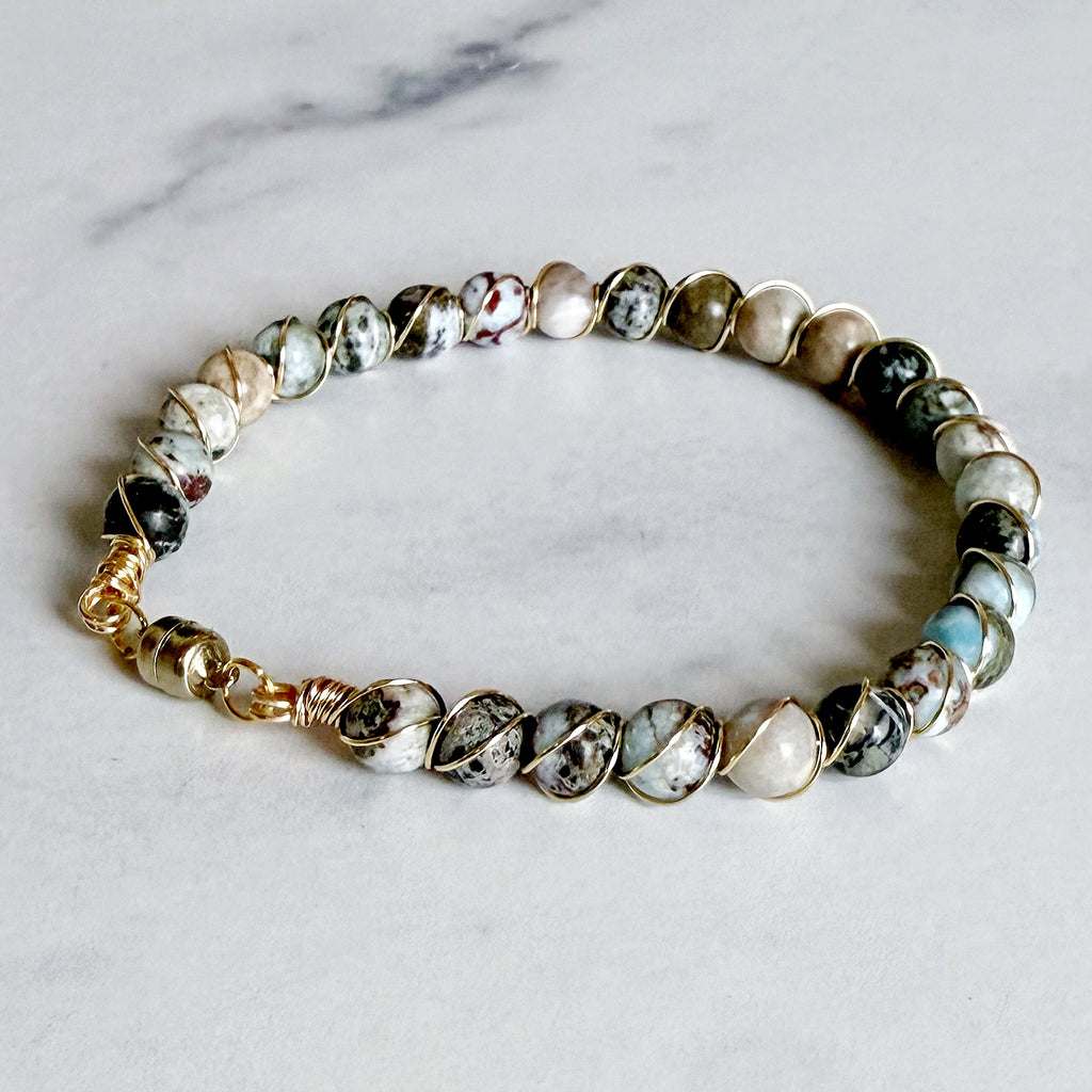 A beaded bracelet featuring natural stone beads in white, black, gray, and light blue, accented with gold-toned metal wire wrapping and a gold clasp, displayed on a light gray marble surface.