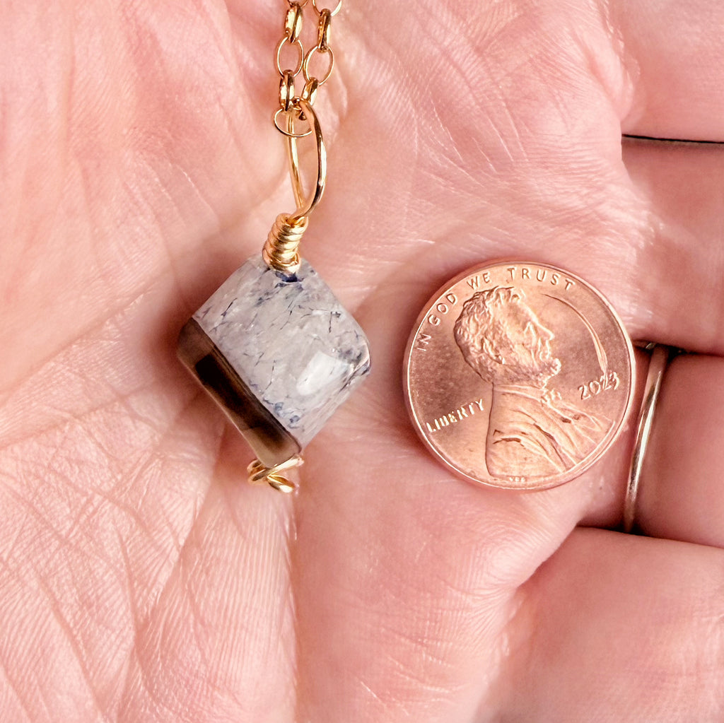 A close-up of a hand holding a small gold chain necklace with a faceted gray stone pendant, shown next to a U.S penny for scale