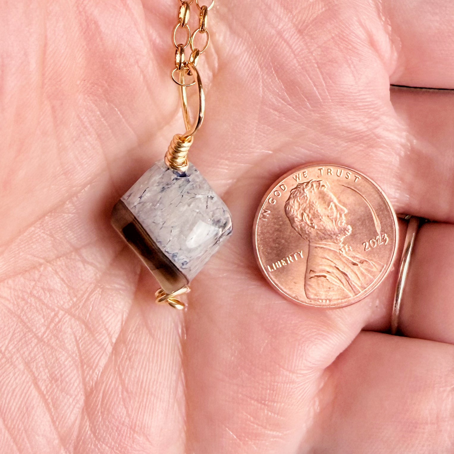 A close-up of a hand holding a small gold chain necklace with a faceted gray stone pendant, shown next to a U.S penny for scale