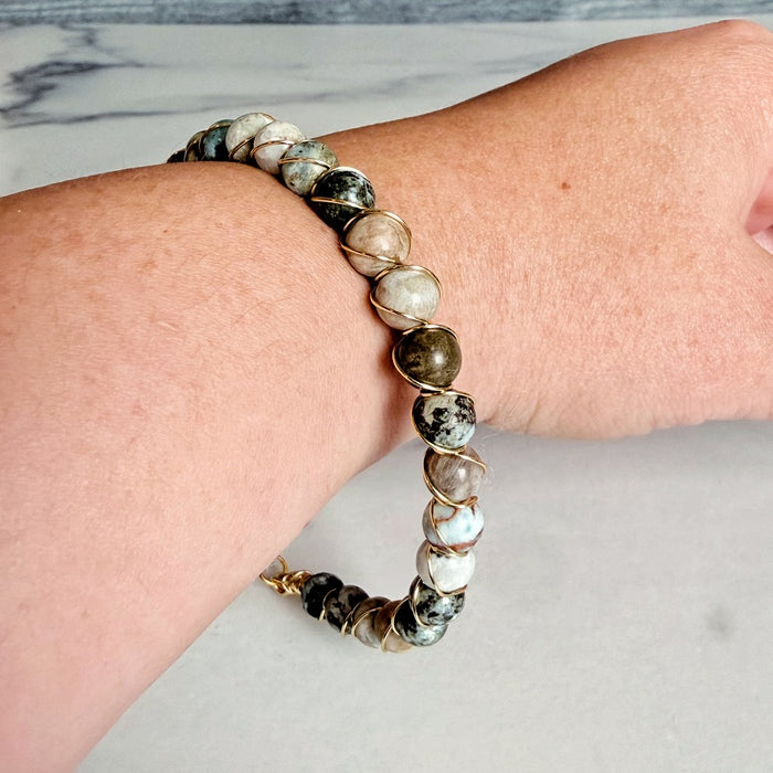 A beaded stretch bracelet with polished round stones in white, gray, black, and olive green, accented by gold-toned wire wrapping, shown worn on a person's wrist.