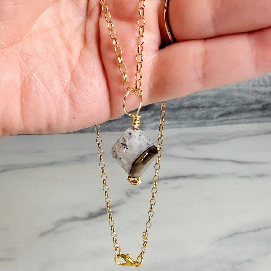 A gold chain necklace with a raw, polished gray and white stone pendant, held in a person's hand against a marble surface background.
