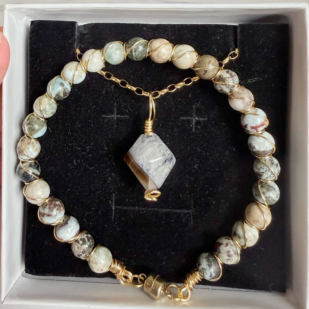 A beaded bracelet with round natural stone beads in green, white, and brown, linked with gold-toned chain and wire-wrapped details, displayed on a black velvet insert inside a white box.