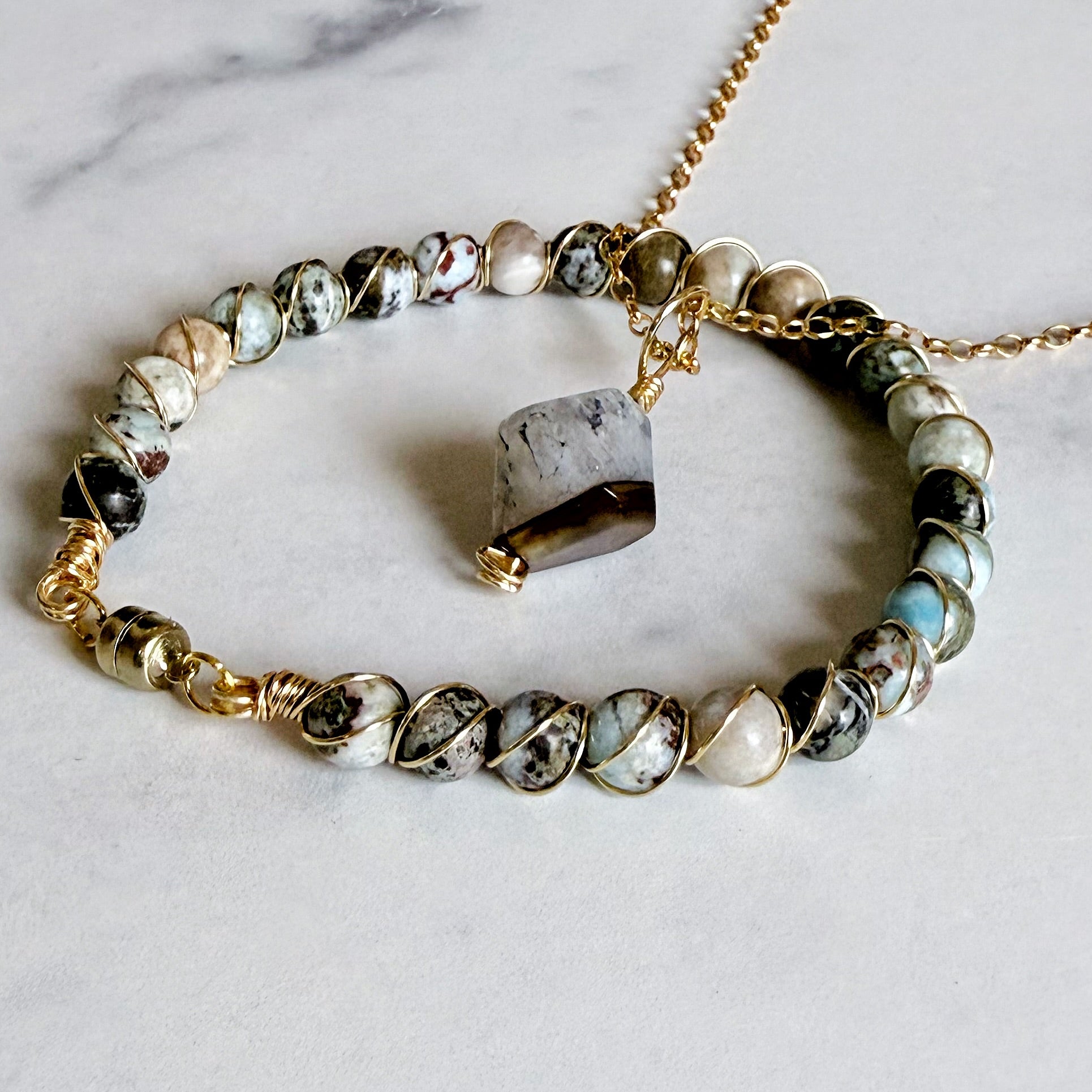 A gold chain bracelet with a beaded stretch section featuring natural stone beads in black, white, and light blue, accented with gold wire wrapping and a central polished gray stone charm, displayed on a light marble surface.