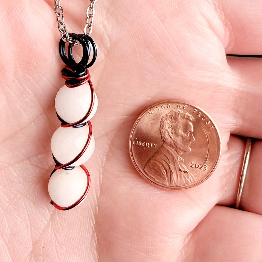 A close-up of a hand holding a silver chain necklace with white oval beads wrapped in black and red wire, shown next to a U.S.