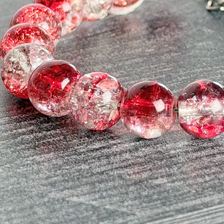 A close-up of a beaded bracelet made of translucent round beads with embedded red and pink glitter, resting on a dark gray textured surface.