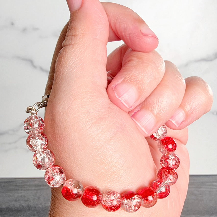 A close-up of a hand wearing a beaded bracelet made of glossy red and clear faceted beads with silver-toned clasp hardware, against a light marble background.
