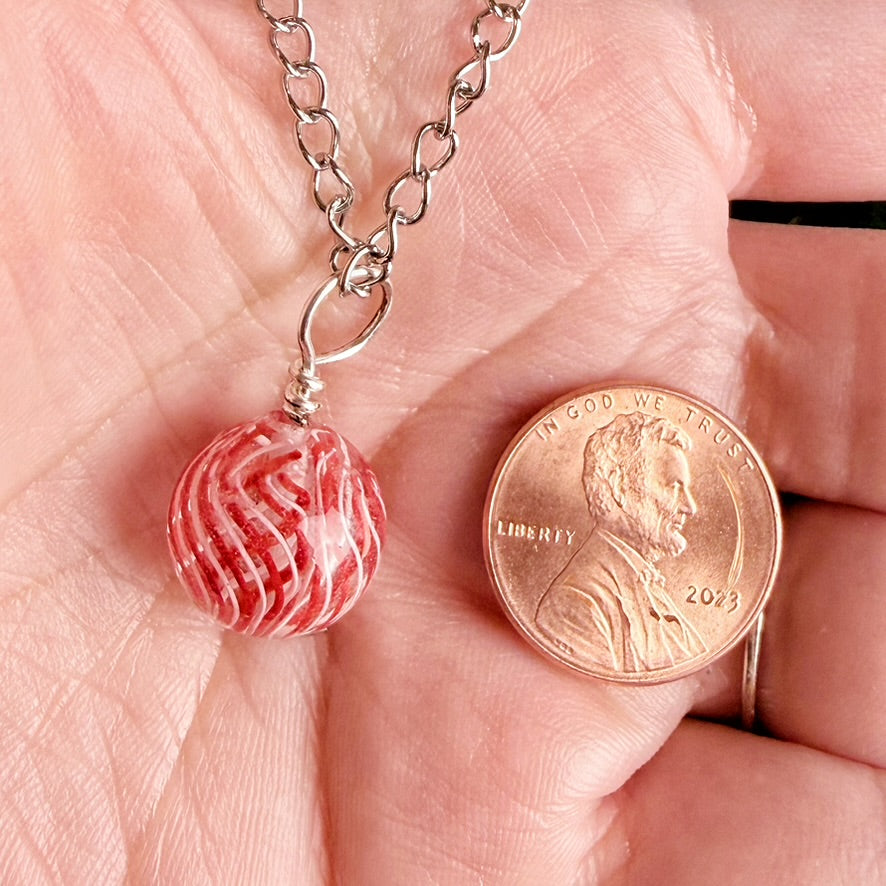 A close-up of a hand holding a silver chain necklace with a red and white marbled bead pendant next to a US penny for scale.