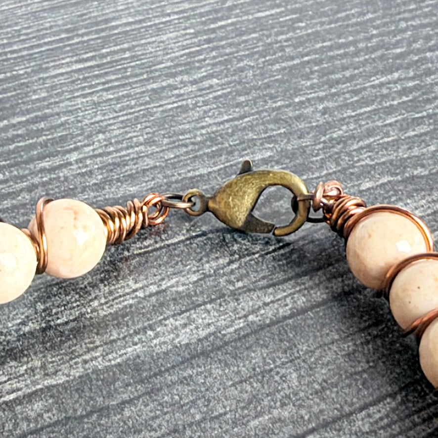 A close-up of a handmade beaded bracelet featuring round cream-colored beads wrapped in copper wire and secured with an antique bronze lobster clasp, displayed on a textured gray background.