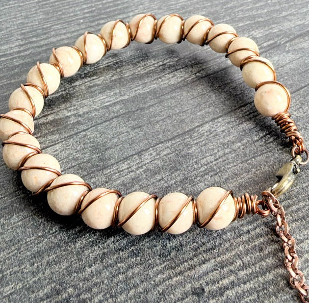 A beaded stretch bracelet made of round cream-colored beads wrapped with copper wire, featuring a small metal clasp and an adjustable copper chain extender, displayed on a gray wood-textured surface.