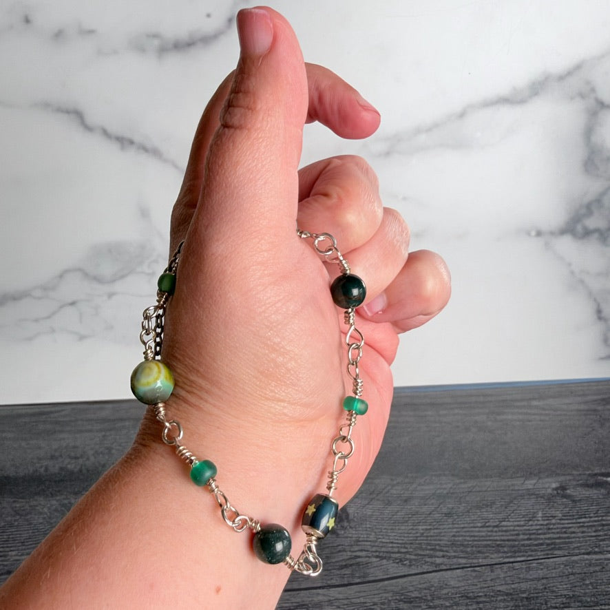 A hand holding a silver chain bracelet with green and black marbled beads, set against a white marble background.