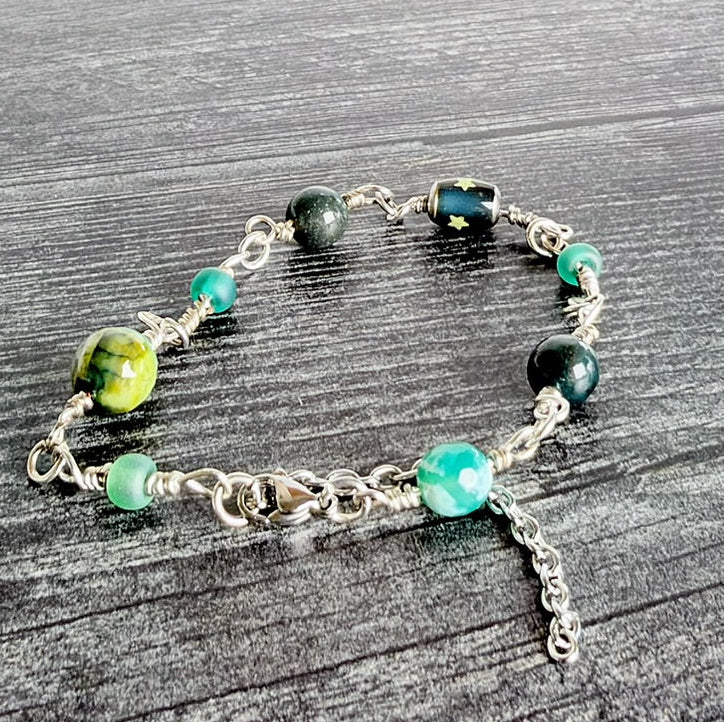 A handmade silver chain bracelet with green and black marbled beads, displayed on a dark gray wood background.