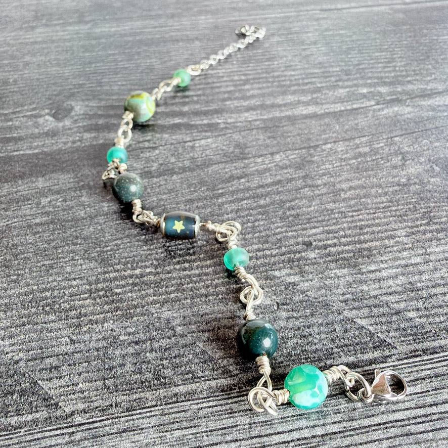 A handmade beaded bracelet with green and teal beads, dark green stone beads, a small star-patterned bead, and silver wire-wrapped chain links, displayed on a gray wood-textured surface.