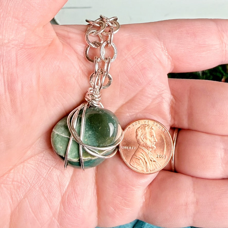 A close-up of a hand holding a silver chain necklace with a wrapped green stone pendant next to a U.S. penny for scale.