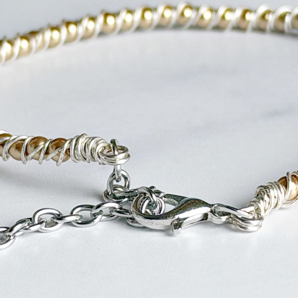 Close-up of a beaded chain bracelet with alternating white and metallic gold beads, wrapped with silver wire at the ends, and secured with a silver lobster clasp on a light gray background.
