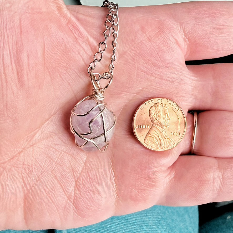 A silver chain necklace with a spherical lavender-pink gemstone wrapped in silver wire, held in a person's hand next to a US penny for scale.