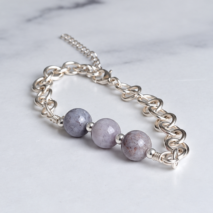 A silver chain bracelet with three round, light purple-gray beads, displayed on a white marble surface.