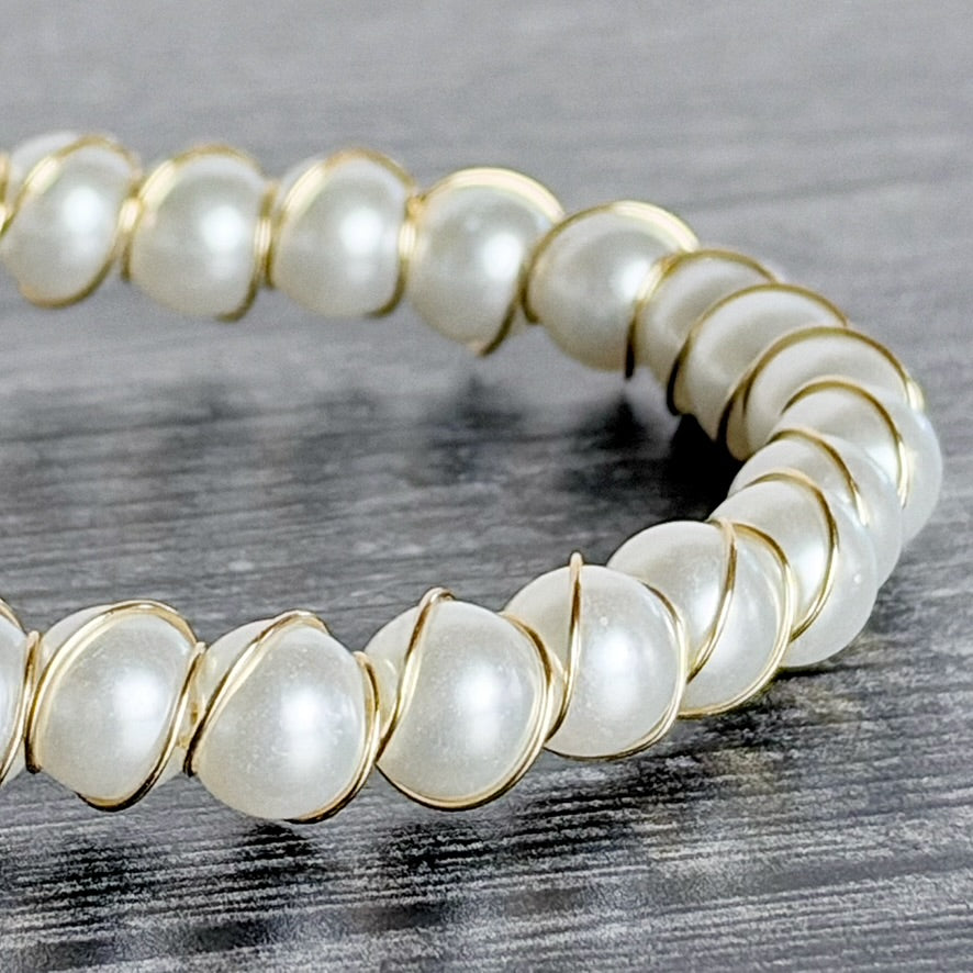 A close-up of a stretch bracelet featuring large, lustrous white faux pearls set in gold-tone metal, displayed on a dark gray wood-textured surface.