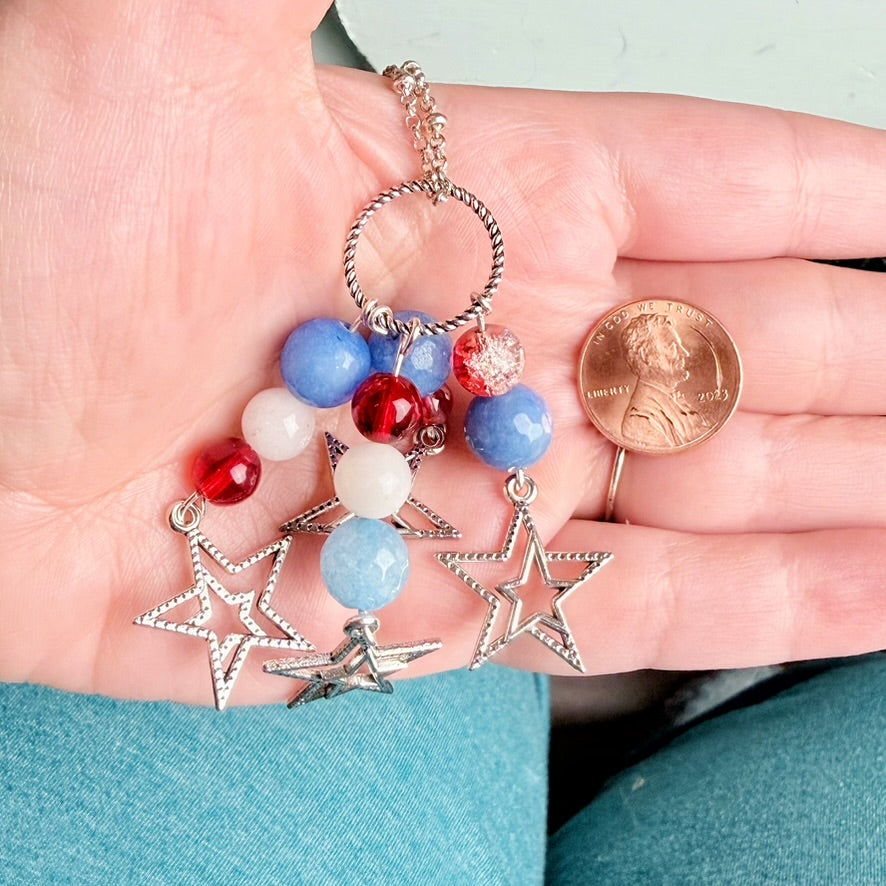 A close-up of a hand holding a patriotic beaded keychain charm with blue, red, and white beads, silver-toned star charms, and a small coin for scale.