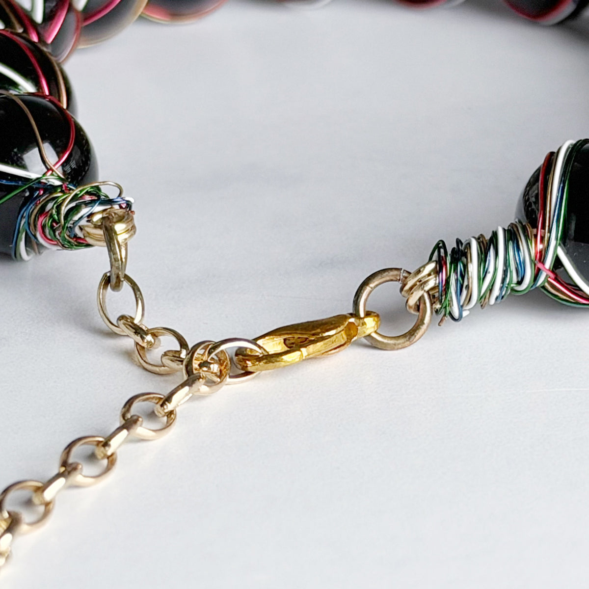 Close-up of a beaded necklace with glossy black round beads wrapped in multicolored wire, a gold lobster clasp, and a gold chain extender on a light gray background.