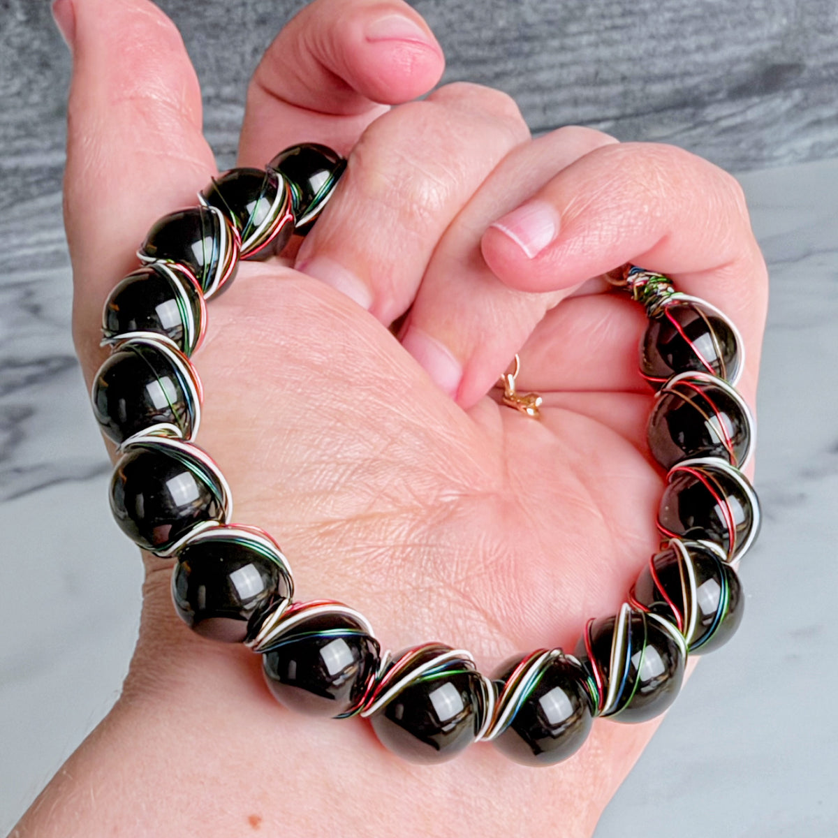 A close-up of a hand wearing a beaded bracelet made of glossy black round beads separated by silver-toned spacers with thin red, white, and green twisted wire details.