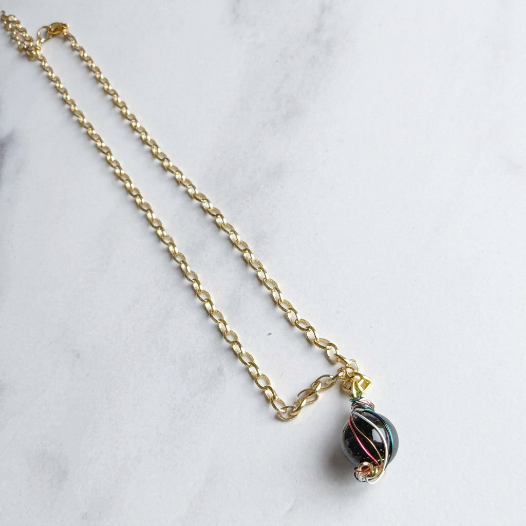 A gold chain necklace with a small gold clasp and a black glass pendant featuring thin red and white stripes, displayed on a light gray marble surface.