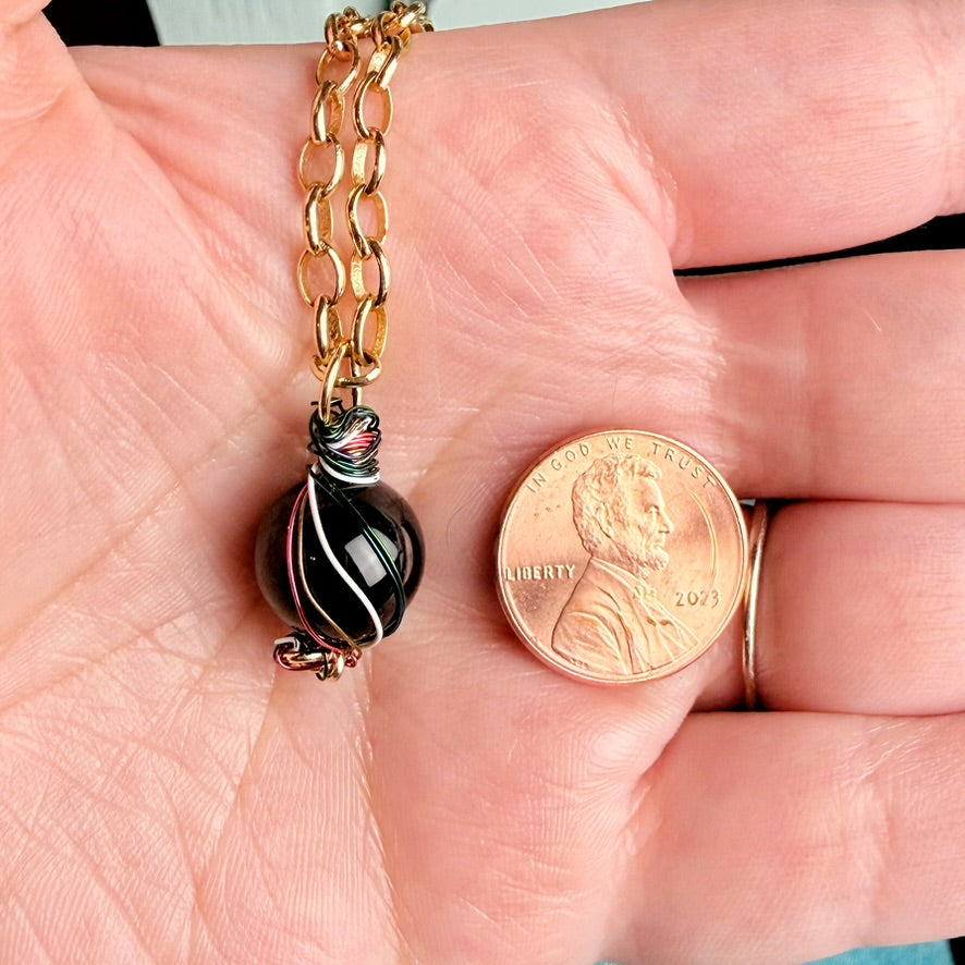 A close-up of a hand holding a gold chain necklace with a black glass bead pendant featuring white and pink swirls, shown next to a US penny for scale.