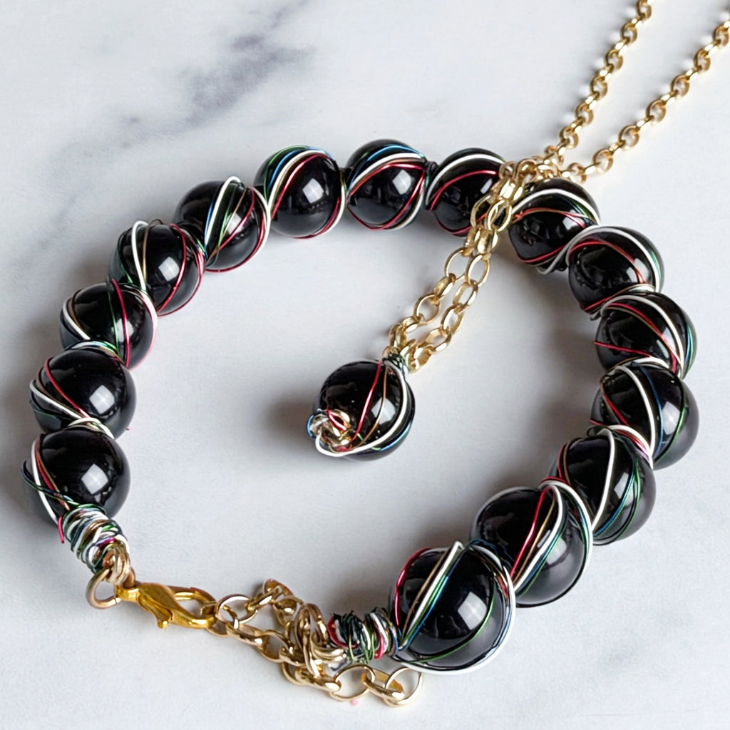 A beaded necklace with glossy black round beads wrapped in silver, red, and green metallic wires, attached to a gold chain and clasp, photographed on a light marble surface.